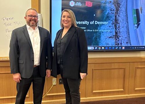 A smiling man and woman standing side by side in front of a large presentation monitor.